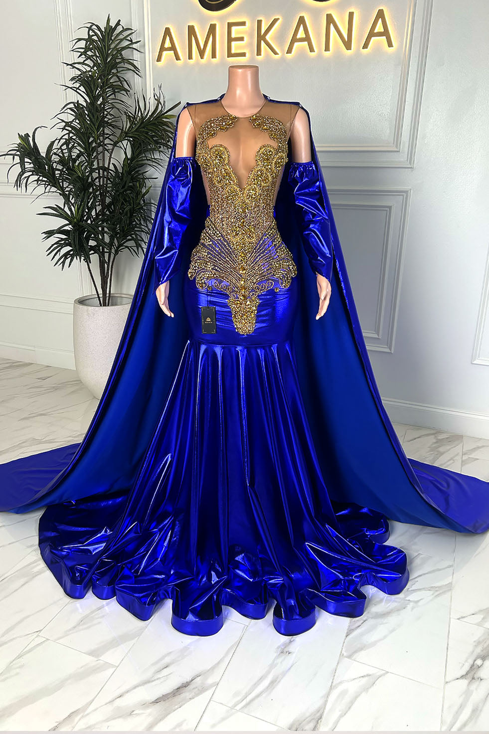 Full length view of electric blue mermaid gown with attached cape and gold embellished illusion bodice, displayed on mannequin under Amekana sign.