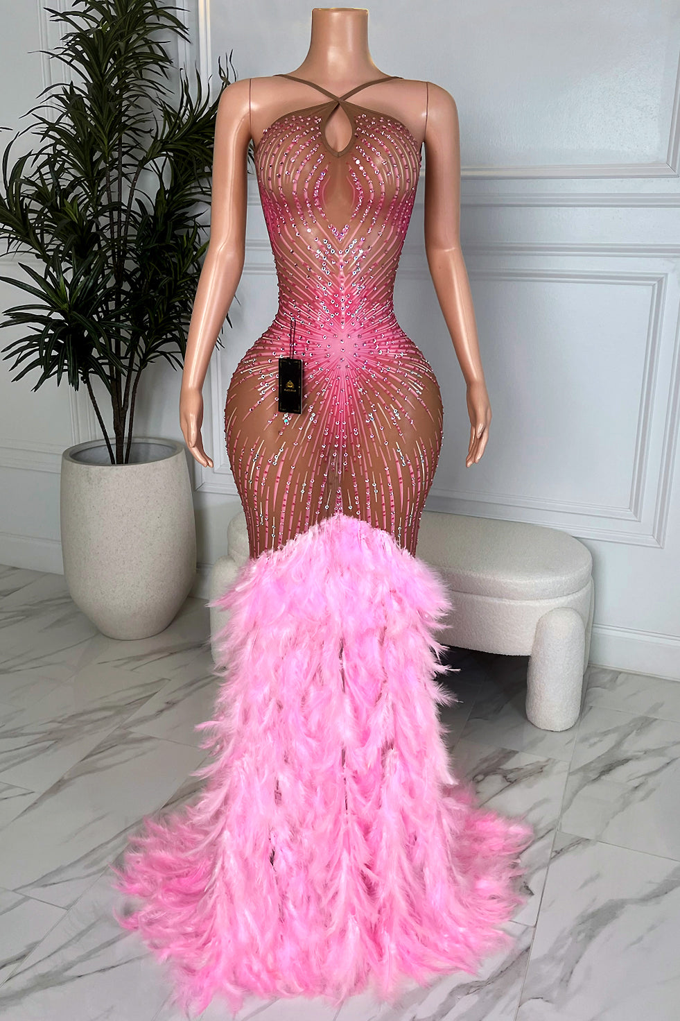 Front view of Amekana Amara Pink Feather Maxi Dress, nude illusion rhinestone gown with pink feather mermaid hem on mannequin in marble room