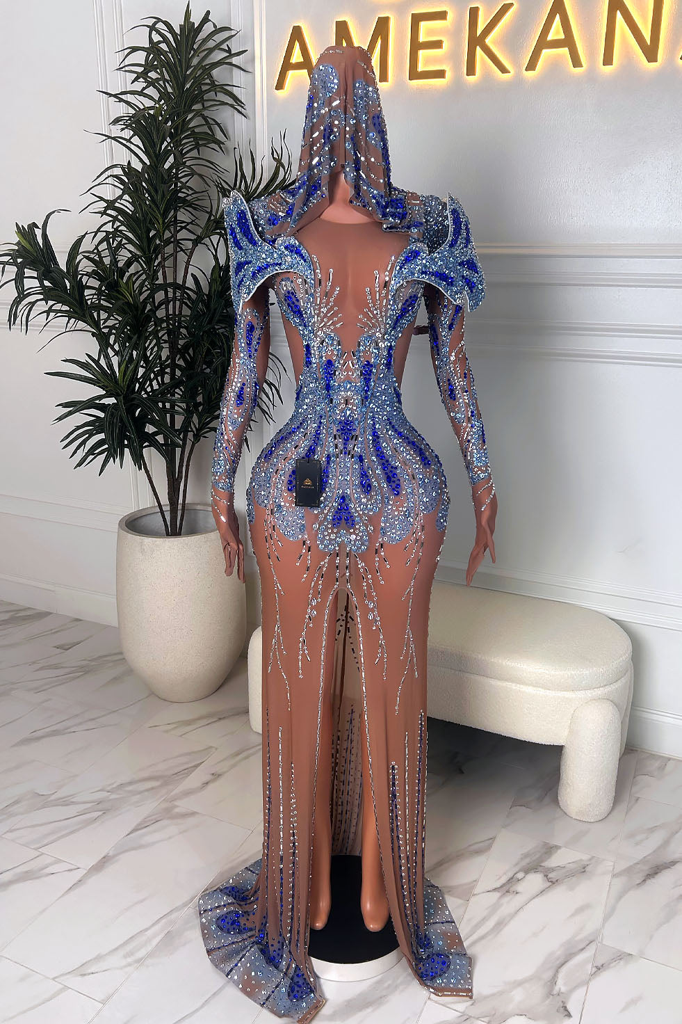 Front view of the same nude illusion gown with the hooded veil worn up, featuring dramatic shoulders, blue rhinestone detailing, and a front slit