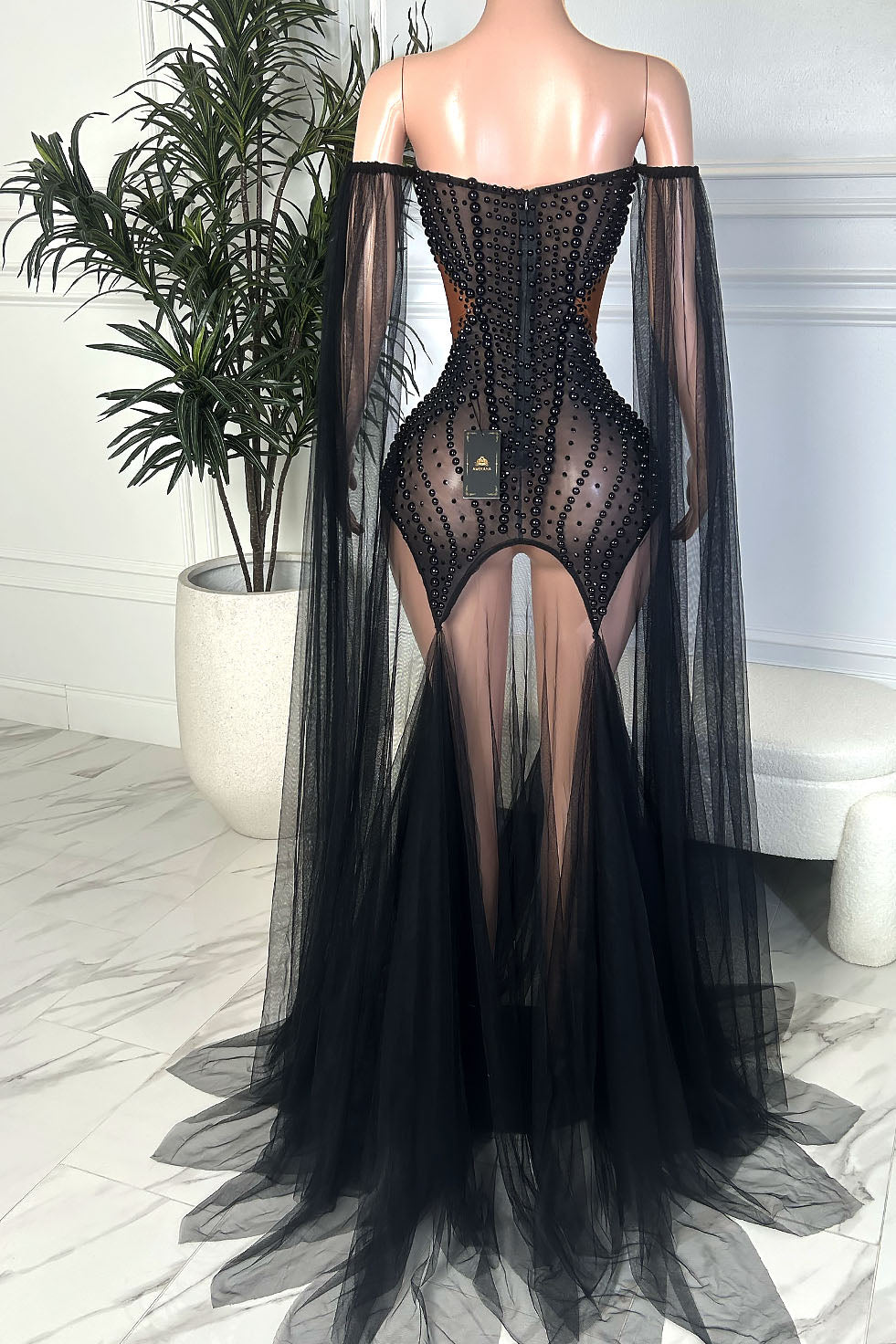 Back view of a black beaded illusion off shoulder gown with sheer mesh, contour bead detailing, off shoulder draped sleeves, and a flowing black tulle train