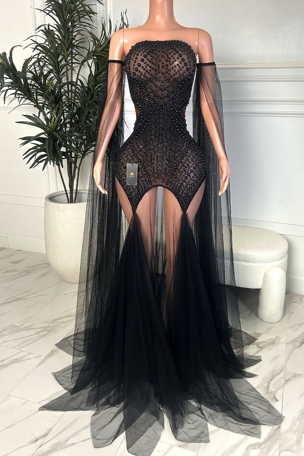 Front view of a black beaded illusion off shoulder gown with sheer mesh, dramatic sleeve drapes, side cutouts, and a flowing black tulle train