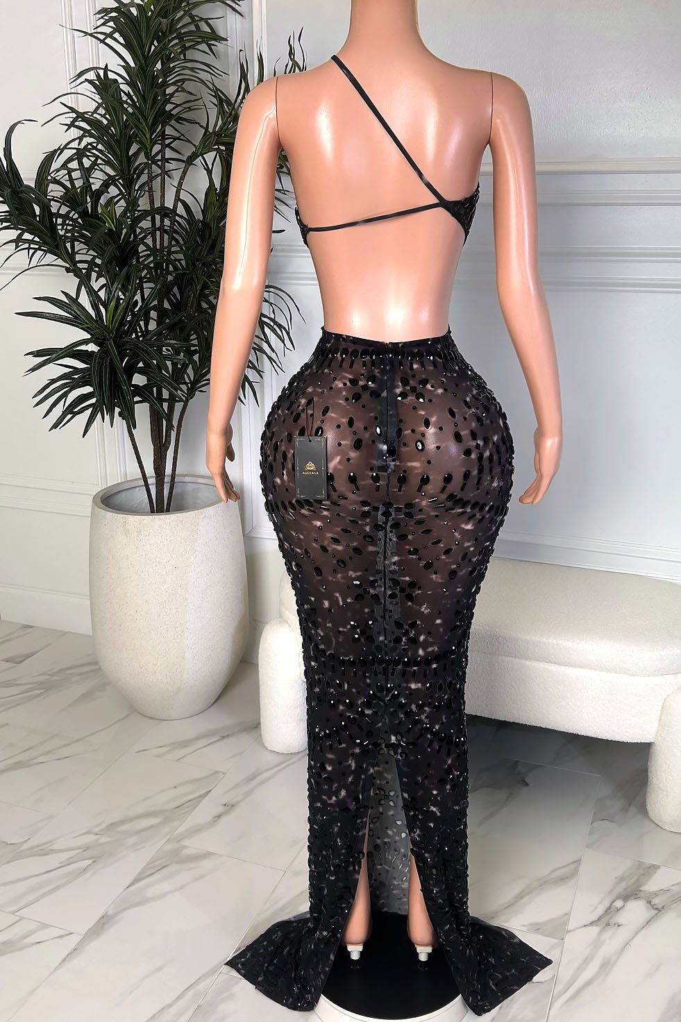 Back view of a black embellished illusion evening gown with an open back, thin asymmetrical strap detail, sheer mesh fabric, and a fitted floor length silhouette with a small train