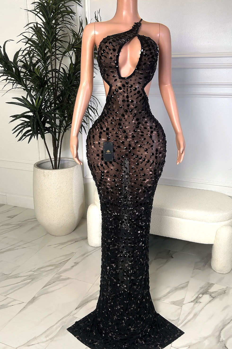Front view of a black embellished illusion evening gown with an asymmetrical neckline, front keyhole cutout, waist side cutouts, and a fitted floor length silhouette