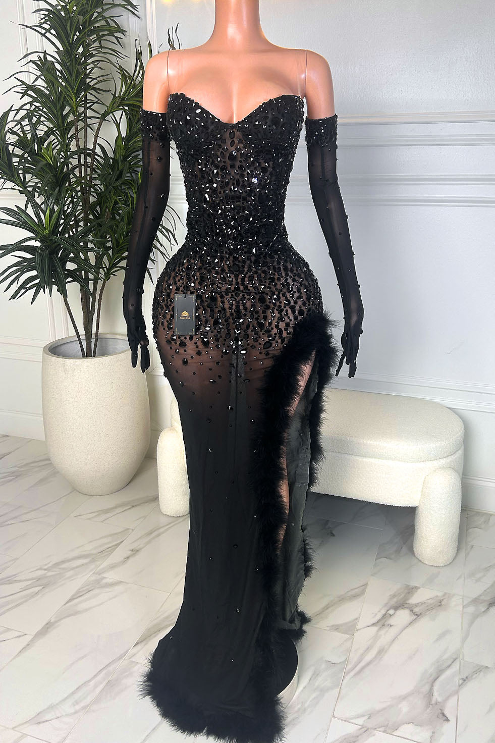 Black off shoulder nude illusion gown with all over black rhinestones, matching sheer glove sleeves, and feather trim along the high slit, front view