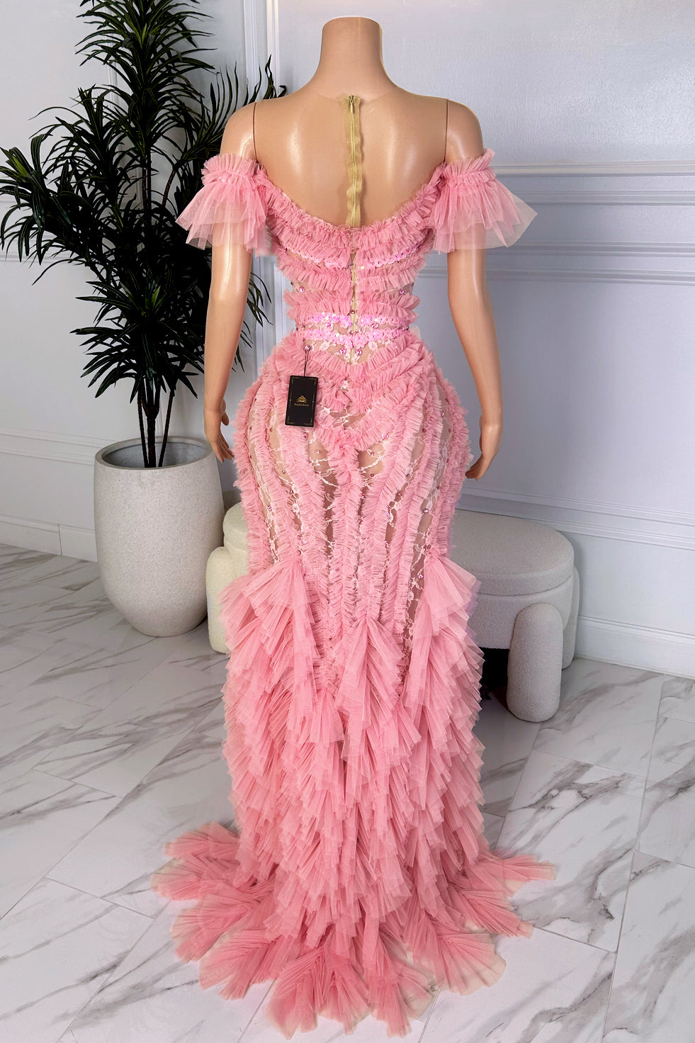 Back view of a soft pink off-shoulder evening dress with layered ruffle detailing, illusion lace panels, and a floor-length hem