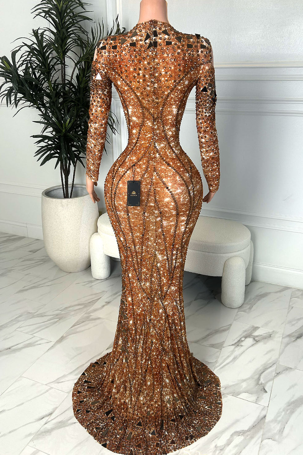 Back view of a bronze mermaid gown with rhinestone and mirror embellishments, long sleeves, fitted silhouette, and sweeping train