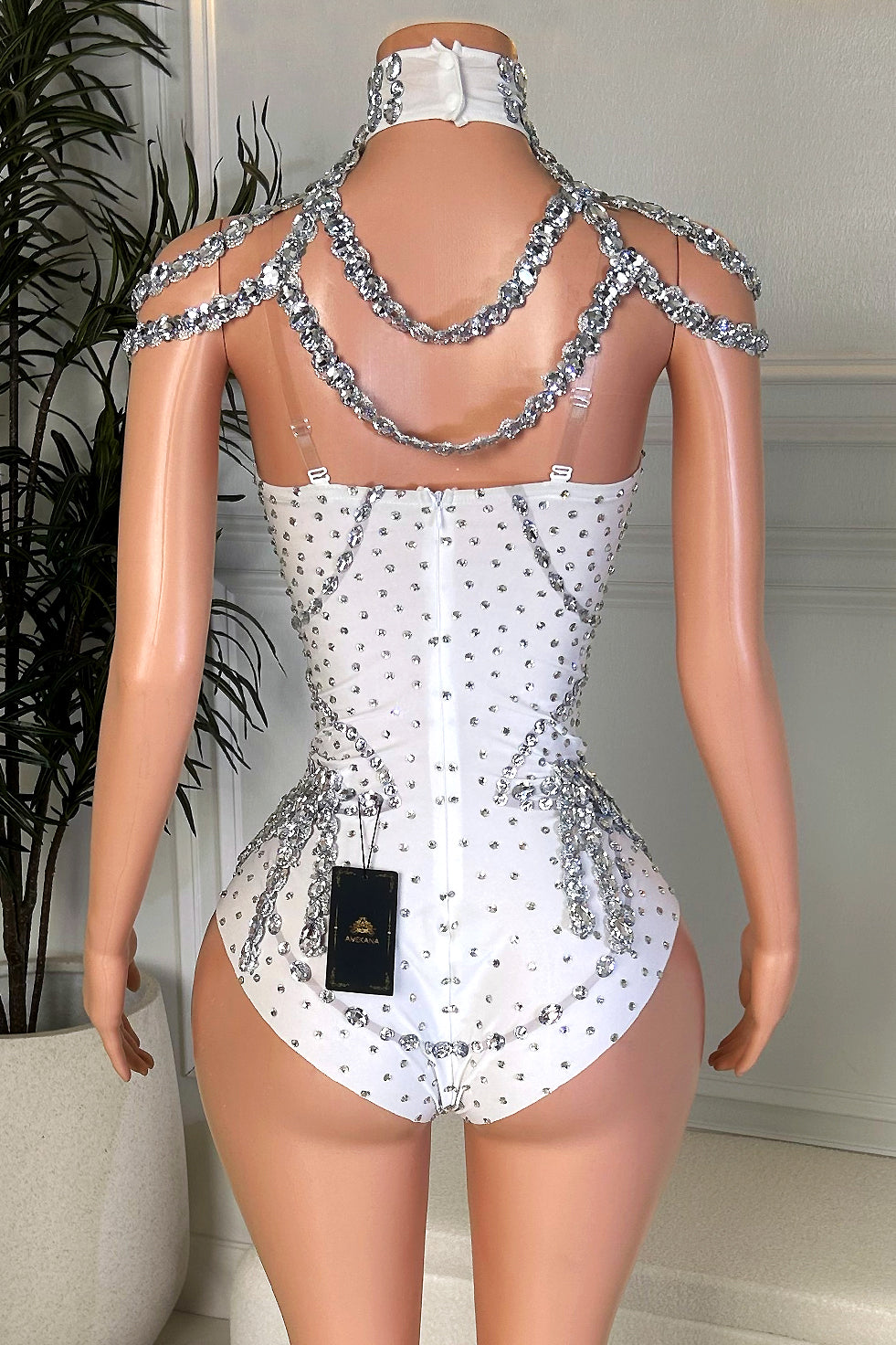 Back view of a white rhinestone playsuit with a jeweled choker neckline, layered crystal drape, clear support straps, and back zipper closure