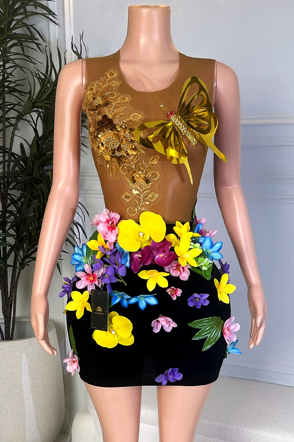 Front view of a brown illusion mesh and black velvet mini dress with gold butterfly appliqué and colorful 3D flowers on the skirt, displayed on a mannequin in a modern showroom.