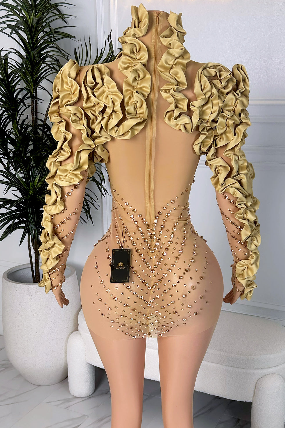 Back view of a gold rhinestone sheer mini dress with high ruffled neckline and long ruffle-trimmed sleeves on a mannequin