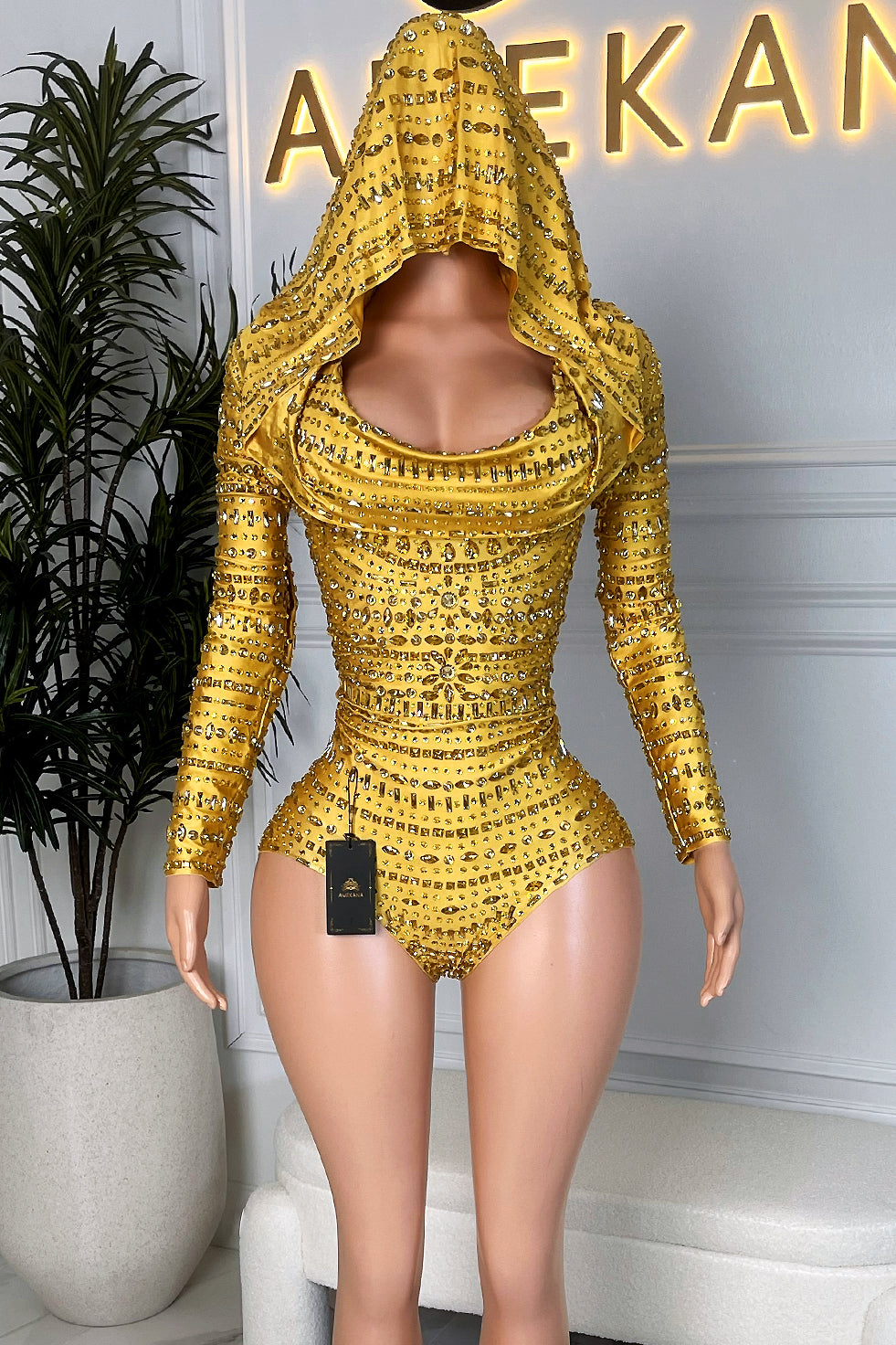 Front view of a yellow rhinestone hooded romper with long sleeves and draped cowl neckline on a mannequin at Amekana”