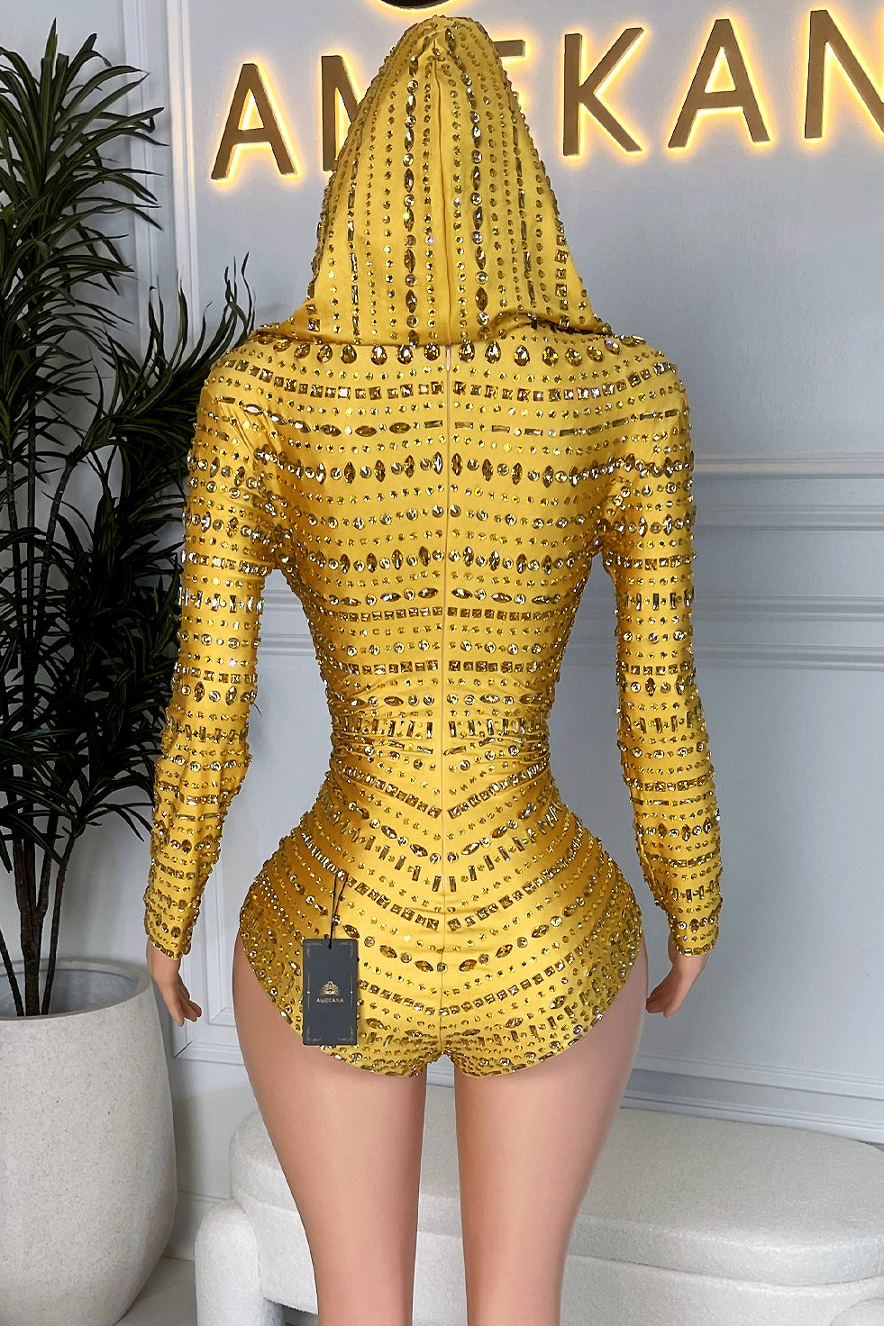 Back view of a yellow rhinestone hooded romper with attached hood and sculpted waist on a mannequin at Amekan