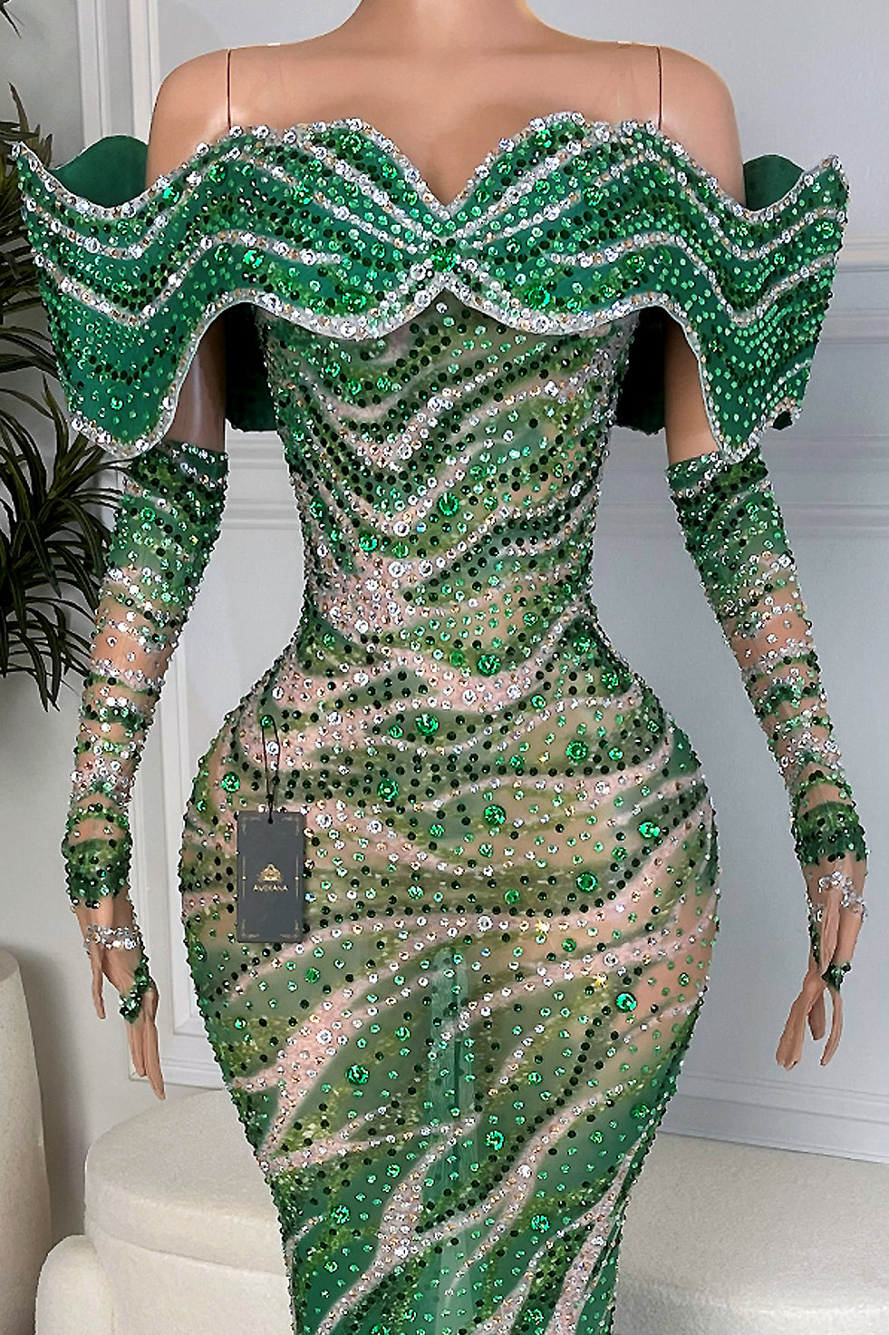 Close-up of emerald and silver rhinestone swirl embellishment on nude mesh with crystal-trim off-shoulder wave ruffle neckline and fitted long sleeves.