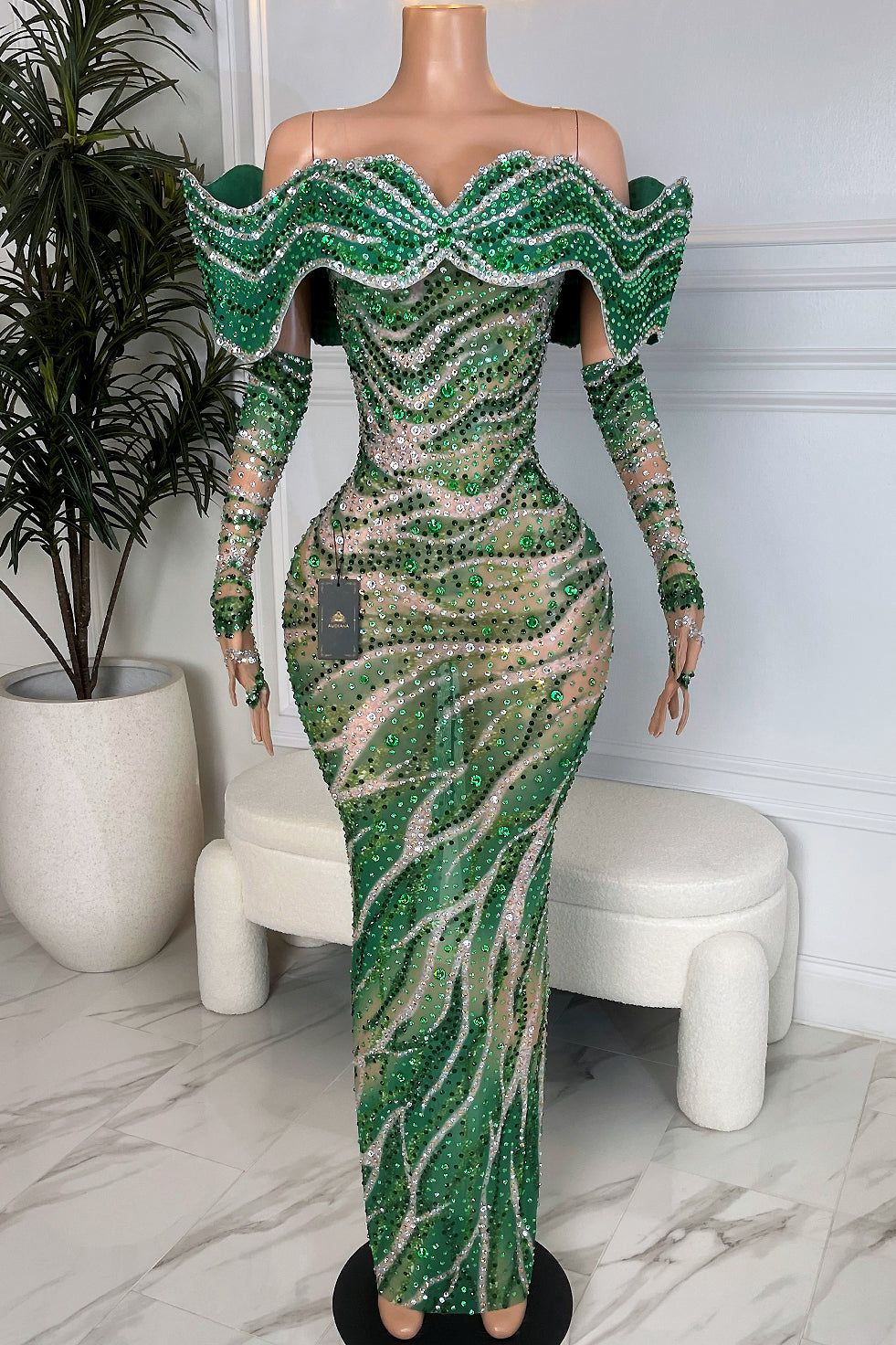 Emerald green rhinestone mesh evening gown with nude illusion base, off-shoulder crystal wave ruffle neckline, long sleeves, and swirling rhinestone pattern.