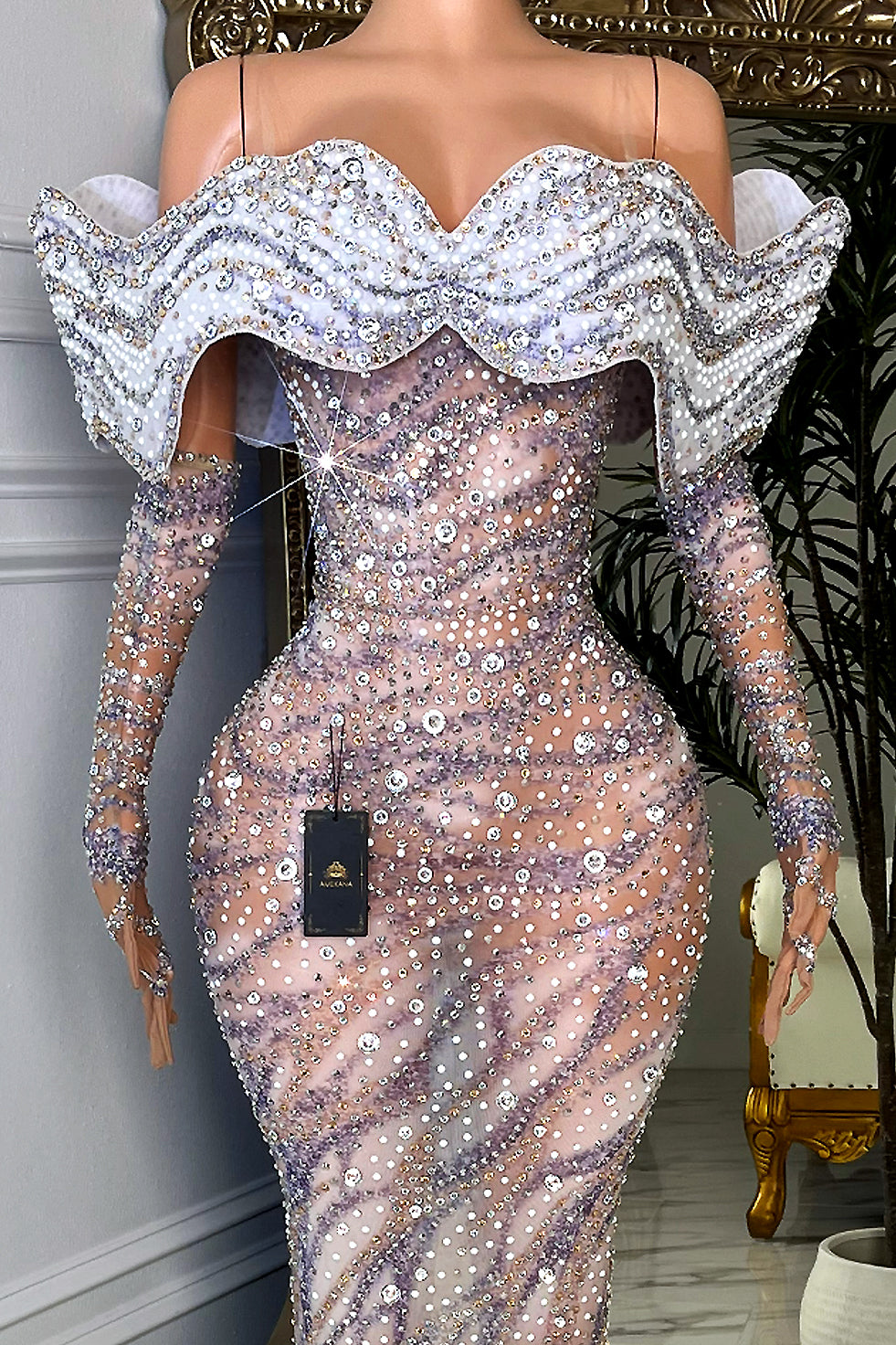 Close-up of nude illusion mesh gown with silver rhinestone scatter, oversized crystal accents, and white off-shoulder wave ruffle neckline trimmed in sparkling stones.