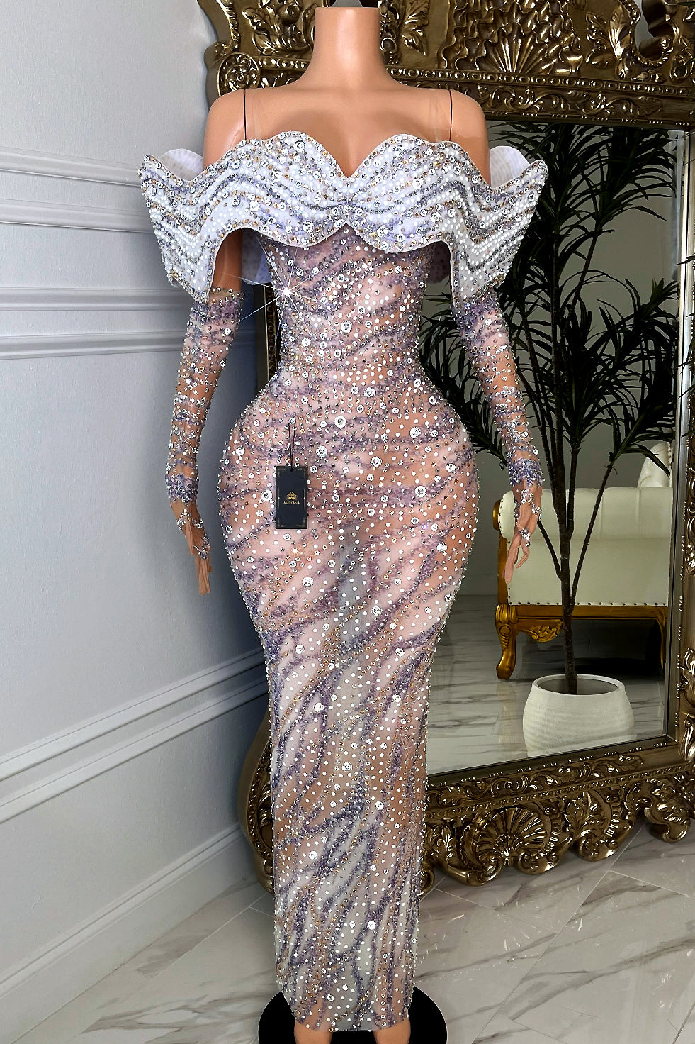 Full-length nude mesh evening gown with silver rhinestone swirl embellishment, long fitted sleeves, and dramatic white off-shoulder crystal wave ruffle neckline.