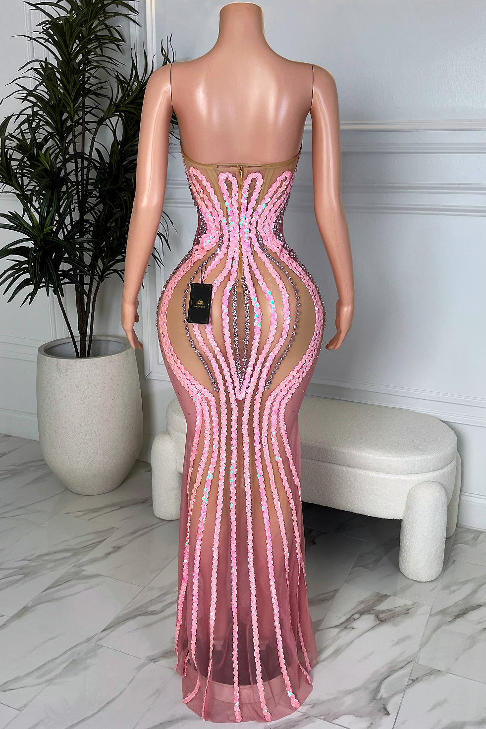 Back view of the Venus Wave Illusion Gown, nude mesh maxi dress with pink and silver sequin contour lines and open back, displayed on a mannequin in a modern showroom.
