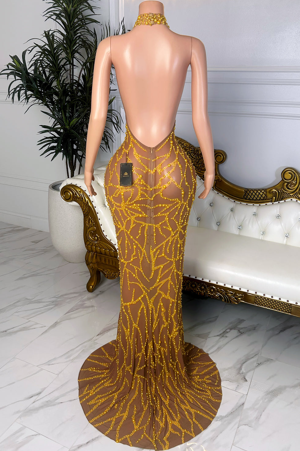 Back view of a nude sheer mesh dress with gold beaded embellishments, high halter neckline, open back, fitted silhouette, and soft trailing hem.