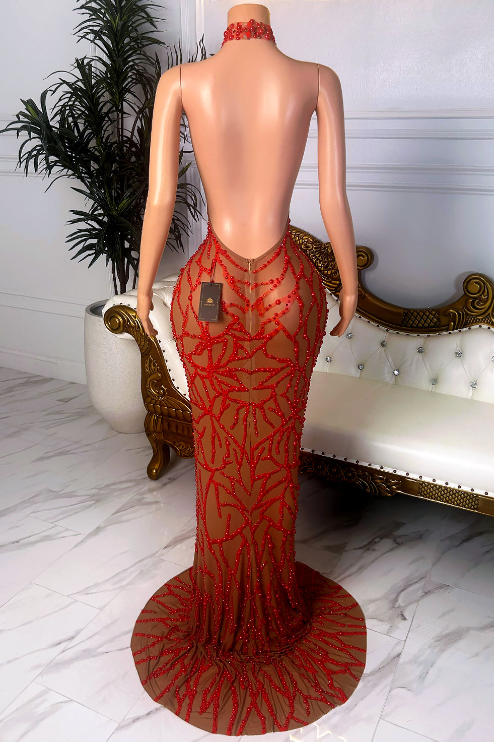 Back view of a nude sheer mesh dress with red beaded floral and vine embellishments, high halter neckline, open back, fitted silhouette, and trailing hem.
