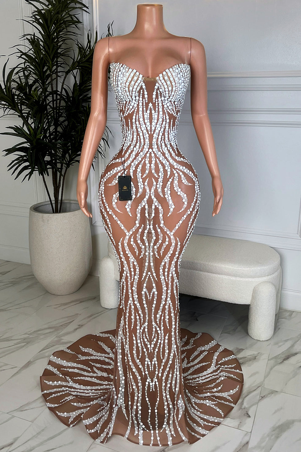 Front view of a nude mesh mermaid gown fully embellished with white rhinestones in wavy flame patterns, strapless sweetheart neckline, and dramatic fishtail train, displayed on a mannequin in a marble floor showroom