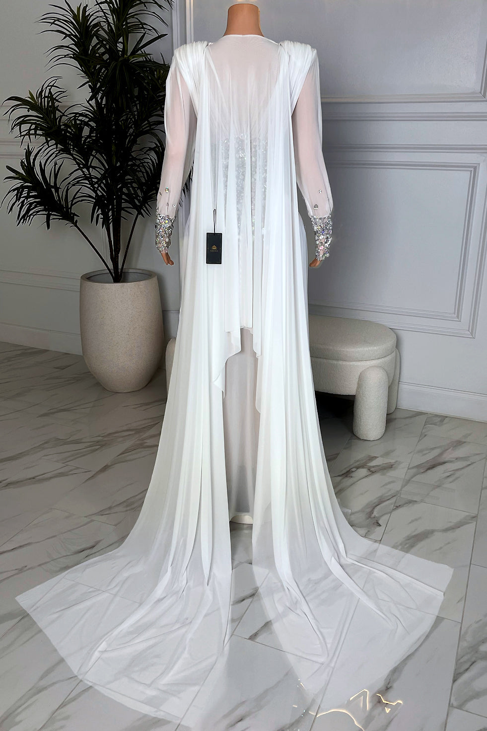 Back view of a long sleeve white sheer rhinestone maxi gown with draped panels, crystal embellished waist, and extended chapel style train from Amekana
