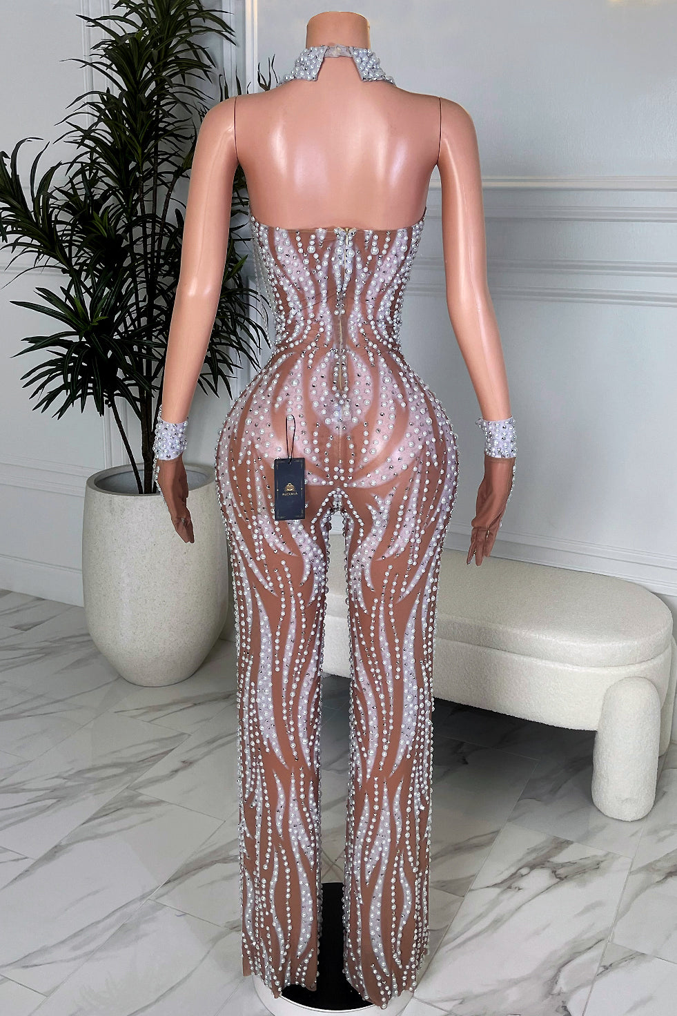 Back view of nude mesh pearl rhinestone jumpsuit with fitted legs, halter neckline, and matching pearl wrist cuffs