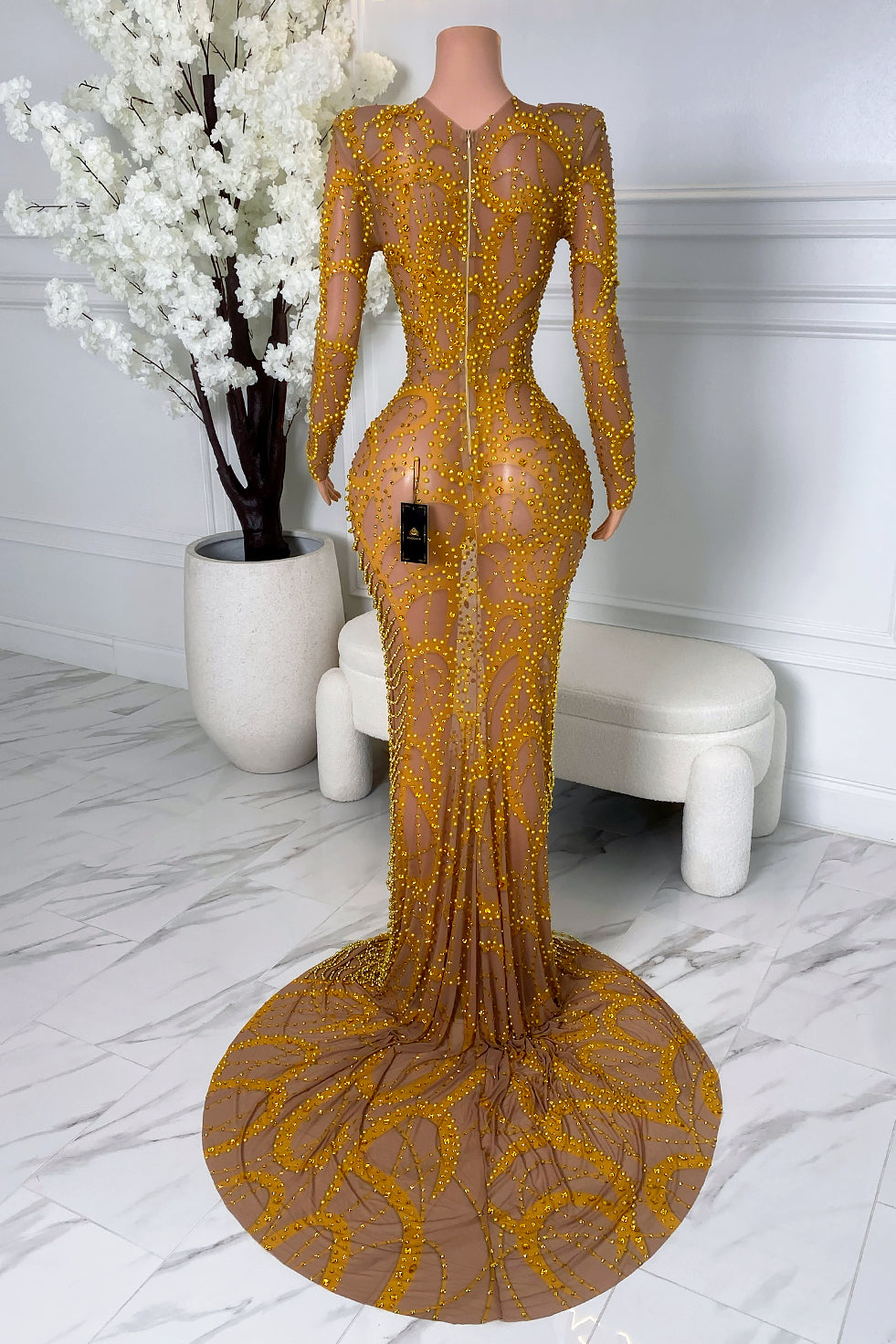 Back view of a nude illusion gold beaded dress with long sleeves, structured shoulders, center back zipper closure, sculpted embellishments, and dramatic circular train